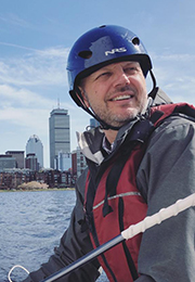 Headshot of David Darmofal wearing a helmet and vest with headphones around his neck and outside on a body of water with a city behind him.