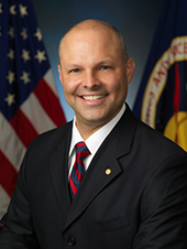 Headshot of Mike Hess wearing a suit and tie smiling with flags behind him