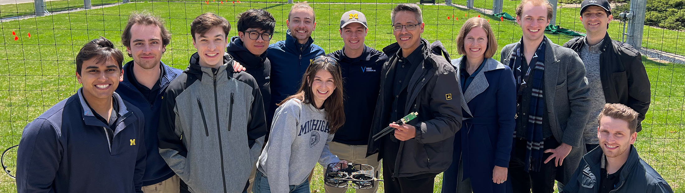 The top half of the M-ACT team standing in front of the M-Air facility with Belcan and Vayu Aerospace representatives holding the M-ACT drone.
