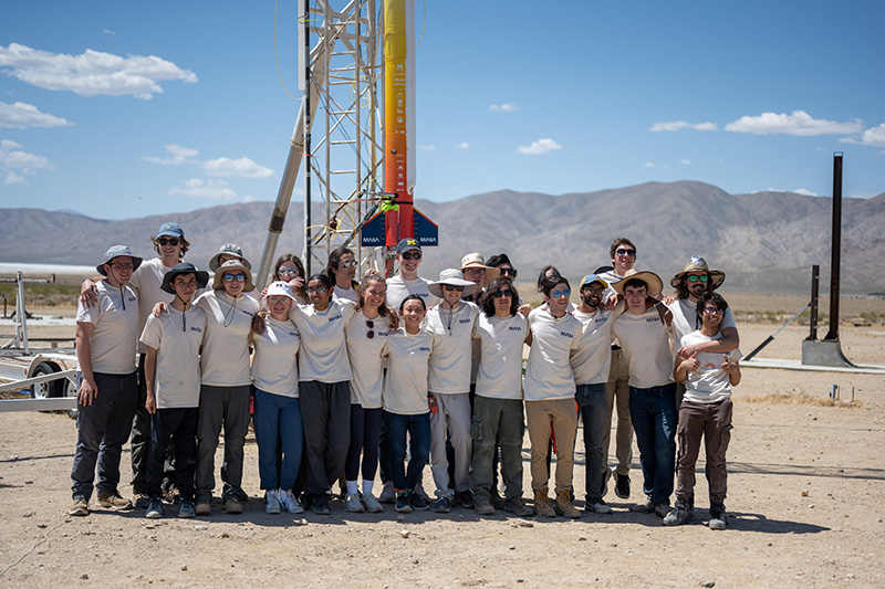 The MASA rocket team all wearing white shirts standing in front of their rocket on the rail posing and smiling in the desert.