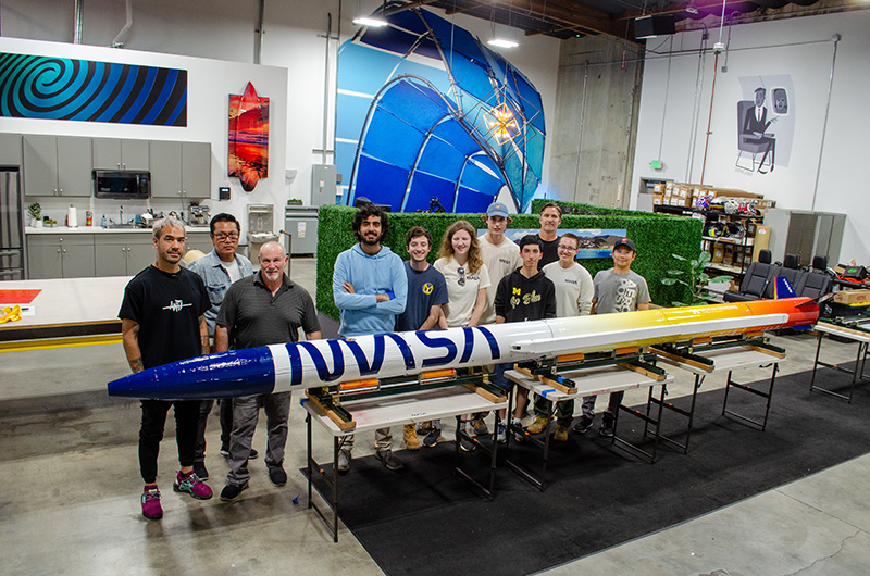 Ten people standing in front of a small rocket inside a warehouse.